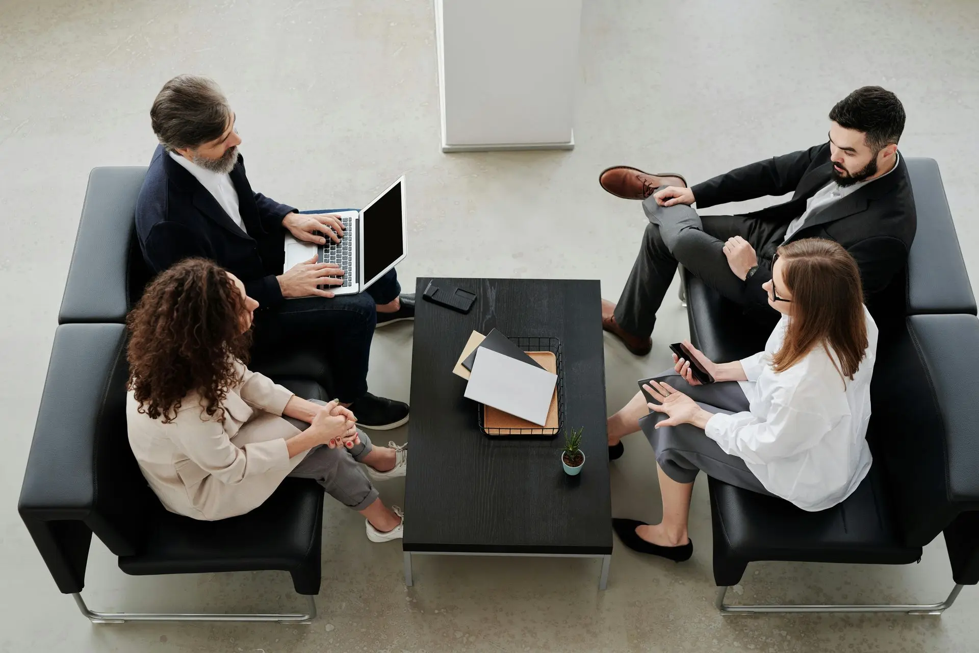 A group of four business professionals having a discussion in a modern office lounge, with laptops, documents, and smartphones on a coffee table.